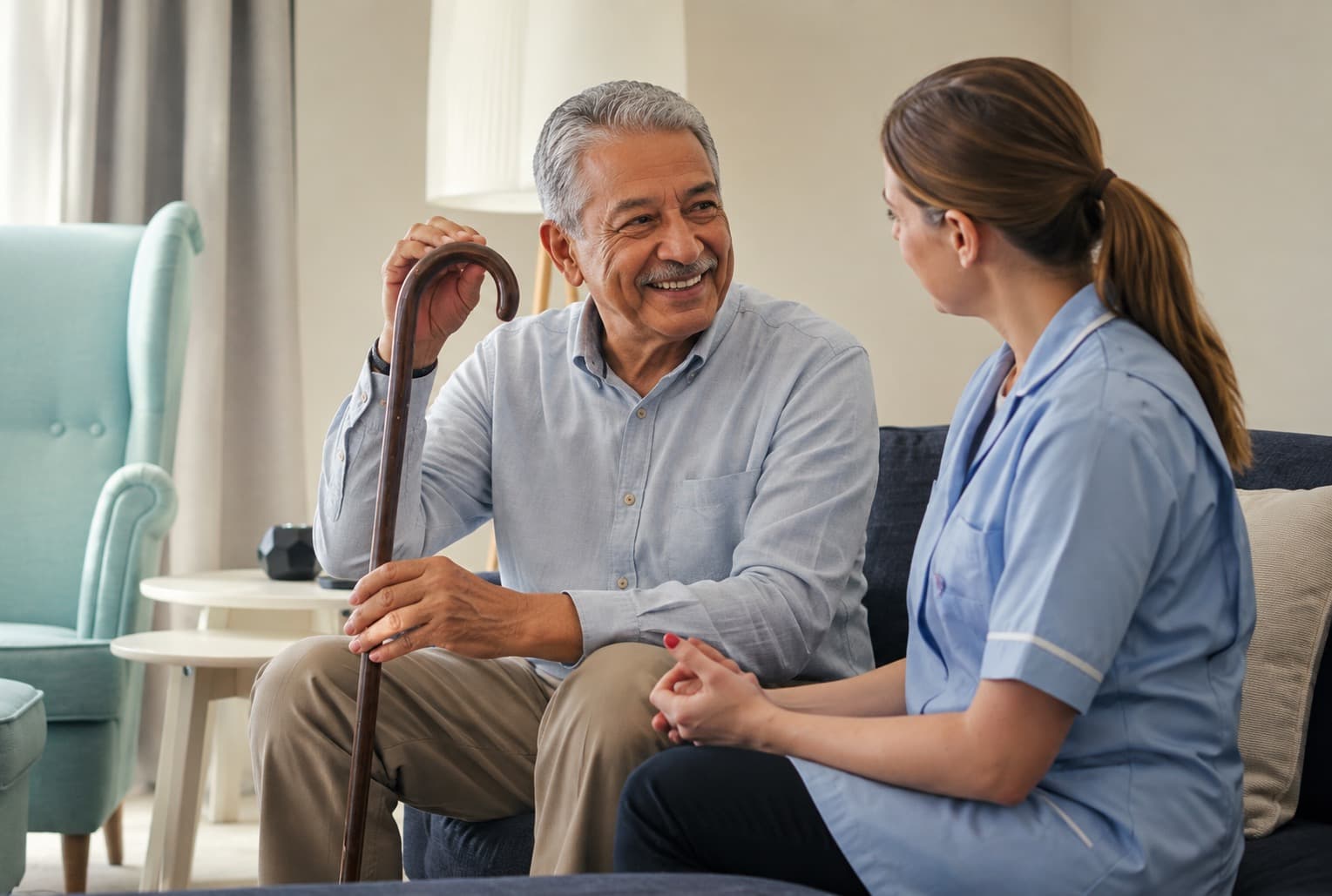 An older man resting on a walking stick in conversation with a care worker on a sofa at home.