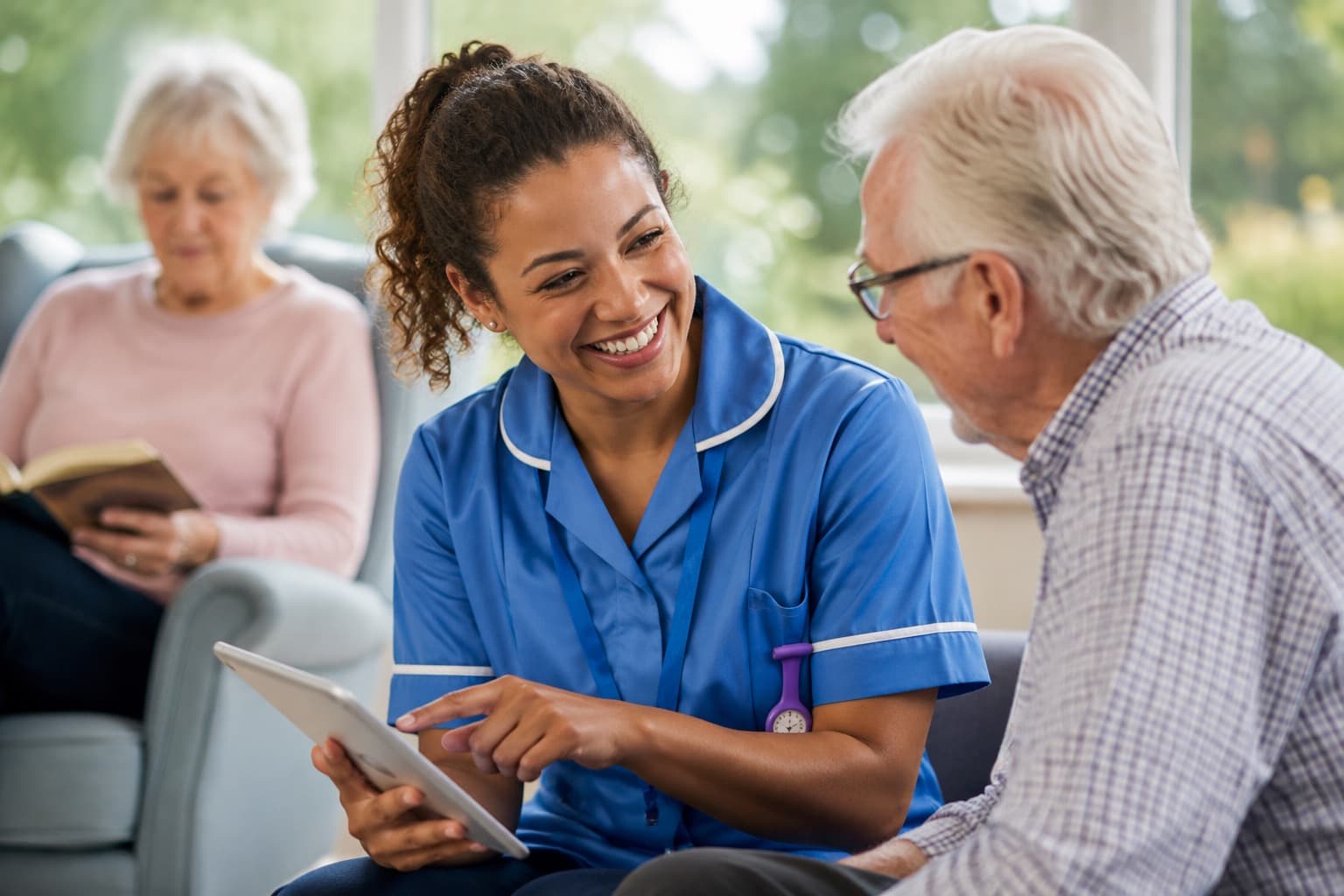 A nurse in uniform showing a tablet to an older man in a bright care home lounge, with another resident reading in the background.
