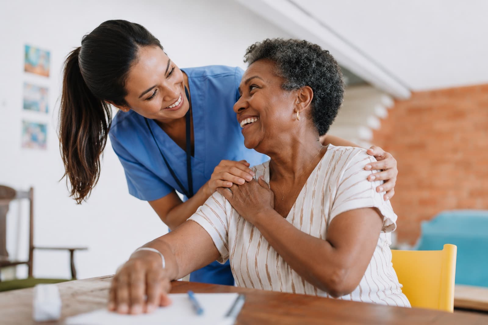 A support worker with an arm around an older woman at a table, both smiling in a bright room.